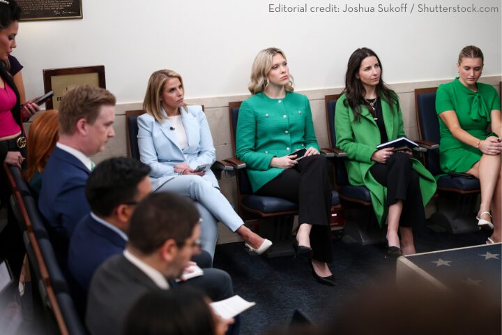 A group of people seated in a conference room, some taking notes. The front row includes two women in green blazers and business attire. The room has formal seating and a professional atmosphere.