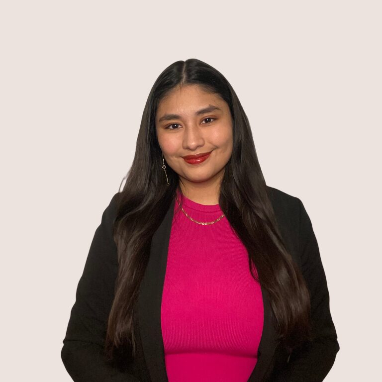 A woman with long dark hair wearing a black blazer over a bright pink top, gold necklace, and earrings, stands against a plain light background, smiling at the camera.