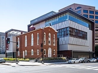 A historic brick church building stands in front of a large, modern glass and metal structure at a city street corner, with cars parked along the curb and a few traffic signals visible.