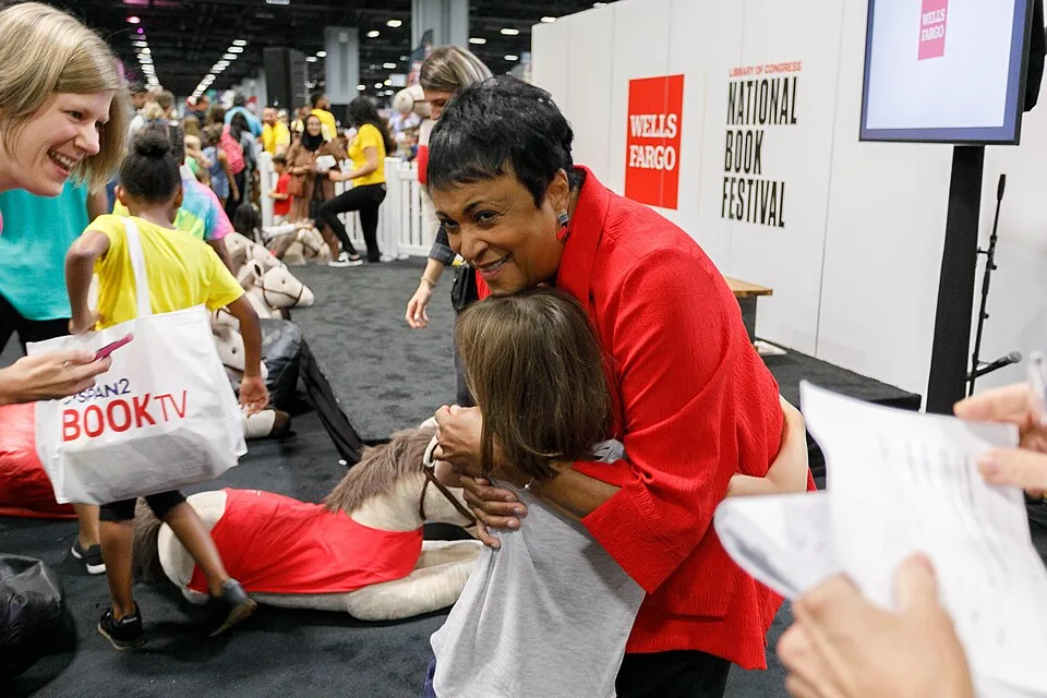 A woman in a red jacket hugs a young child at a crowded National Book Festival event. Other people stand nearby, some holding bags, with festival signage and decorations visible in the background.