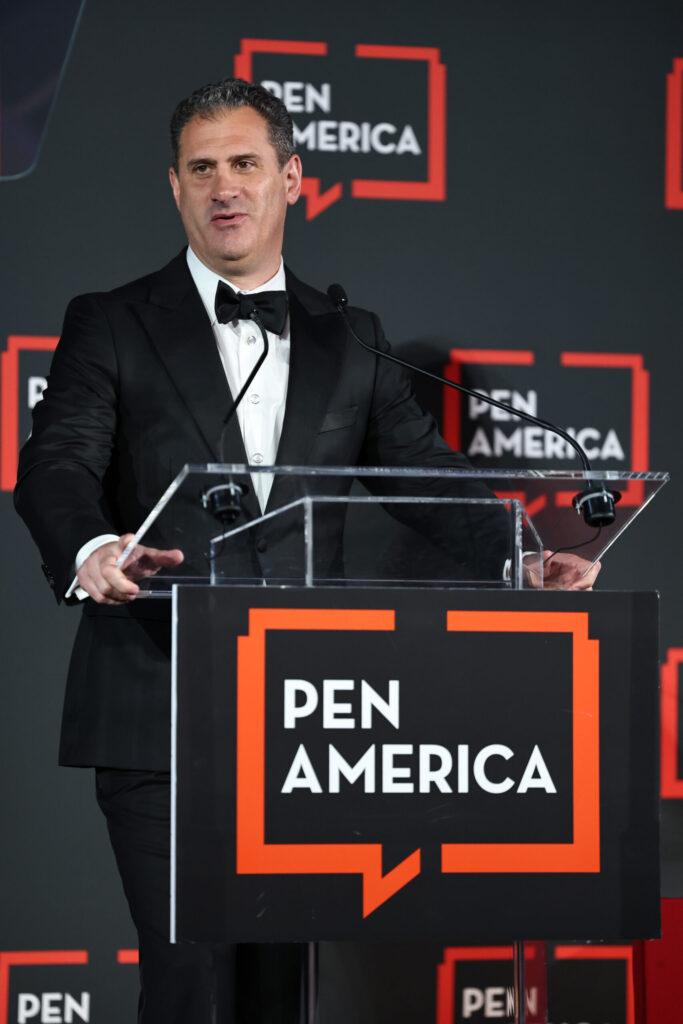 A man in a black tuxedo speaks at a podium with a microphone, featuring the PEN America logo, against a backdrop with repeated PEN America branding.