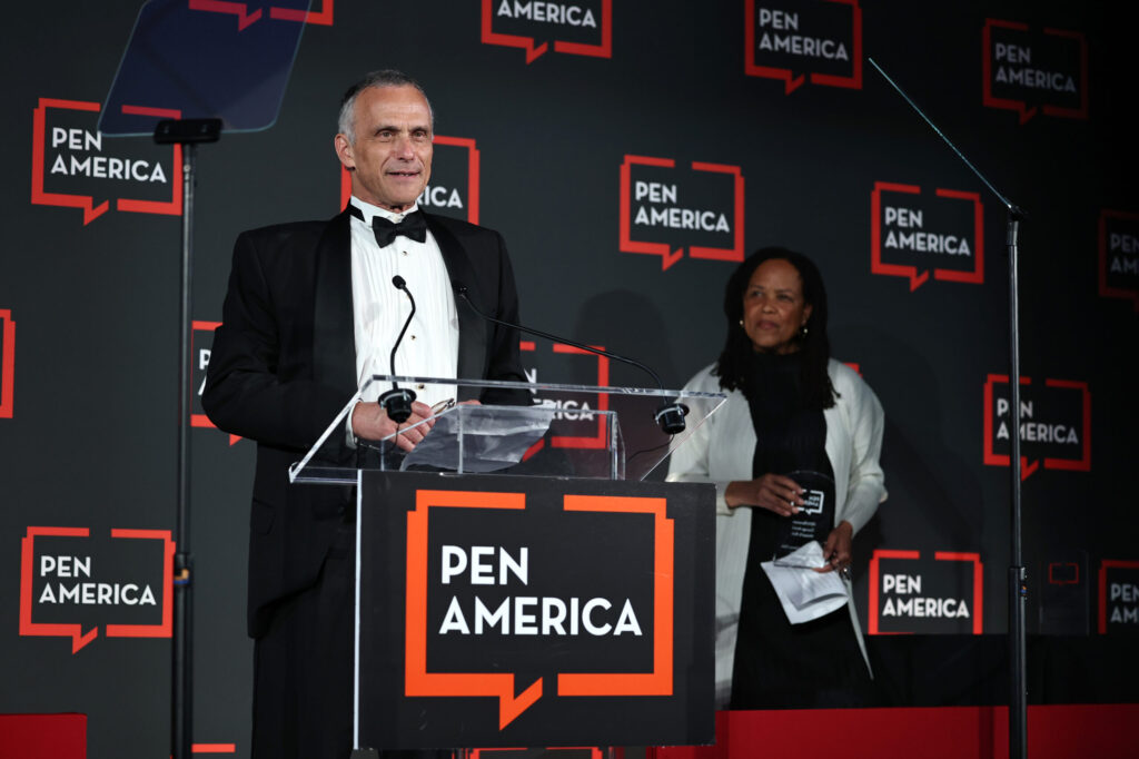 A man in a tuxedo speaks at a clear podium with a “PEN America” sign, while a woman stands nearby holding papers. The backdrop displays multiple “PEN America” logos.