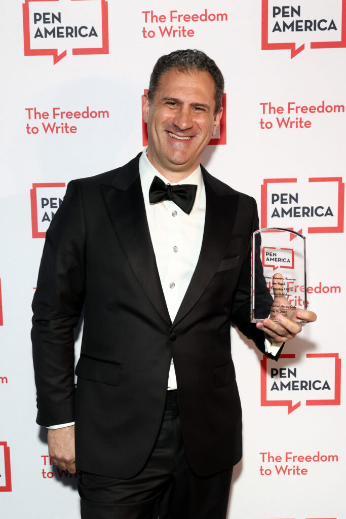 A man in a black tuxedo smiles while holding a PEN America award trophy. The backdrop behind him reads PEN America and The Freedom to Write in red and white.