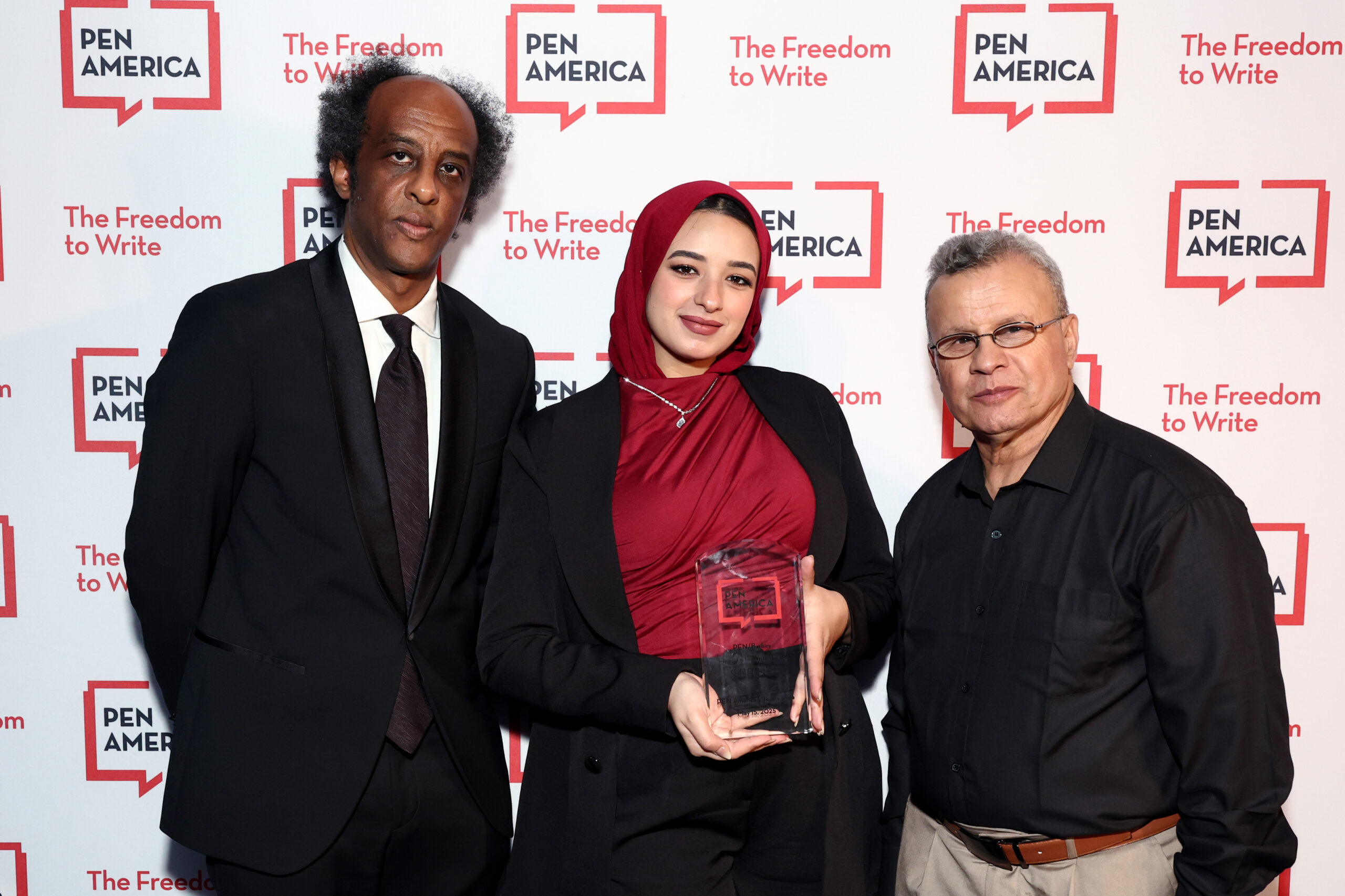 Three people stand together at a PEN America event. The person in the center holds a clear award and wears a red hijab and black jacket. The other two people stand on either side, dressed in formal attire, in front of a PEN America backdrop.