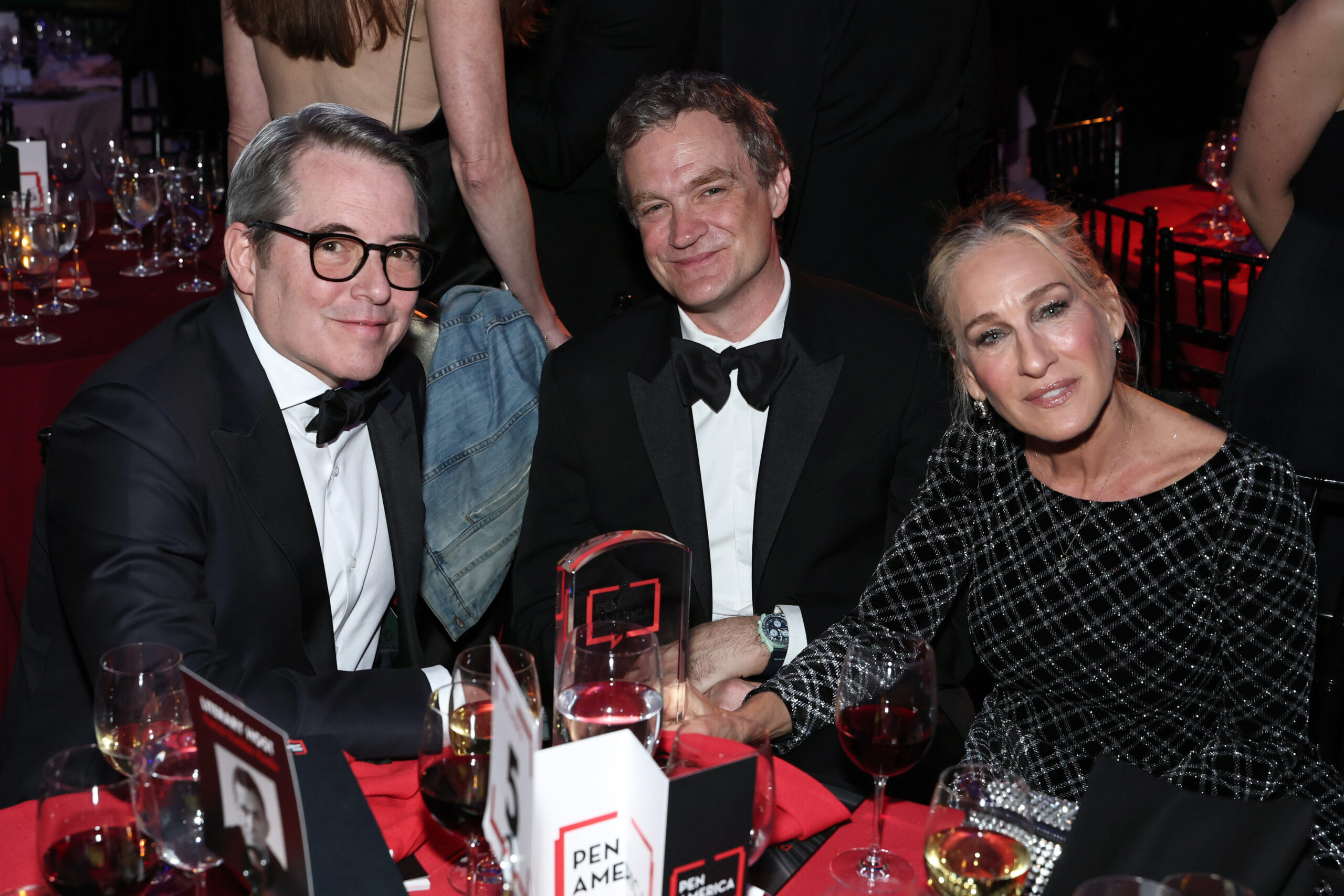 Three people dressed in formal evening wear sit at a red-covered table at an event, smiling at the camera, with glasses of wine and an award visible on the table in front of them.