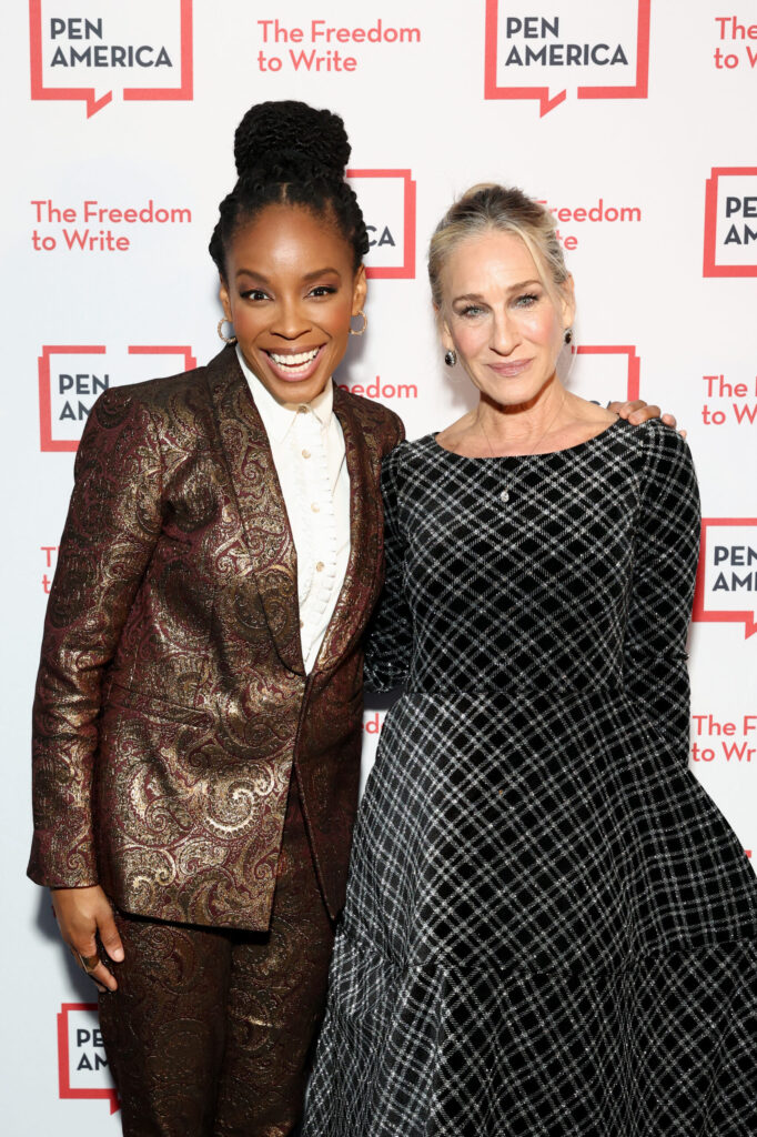 Two women pose and smile together at a PEN America event. One wears a patterned brown suit and the other wears a black and silver checkered dress. The background features PEN America and The Freedom to Write logos.