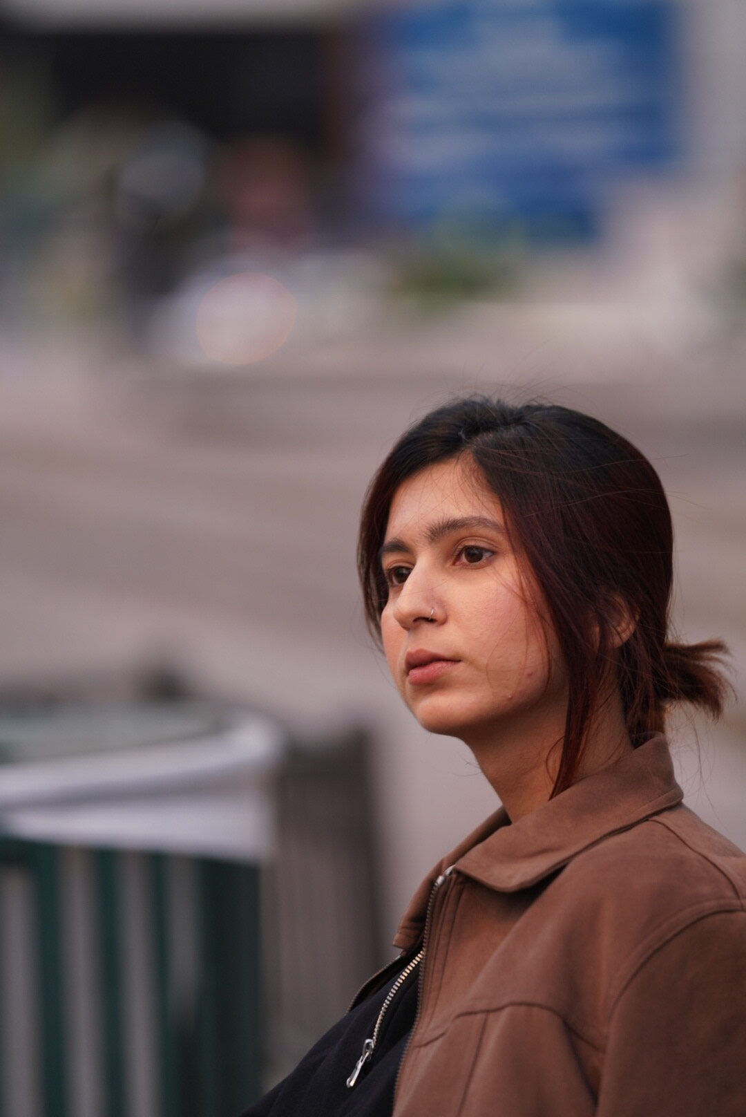 A young woman with long dark hair tied back, wearing a brown jacket, looks thoughtfully into the distance. The background is blurred, giving a soft, urban feel.