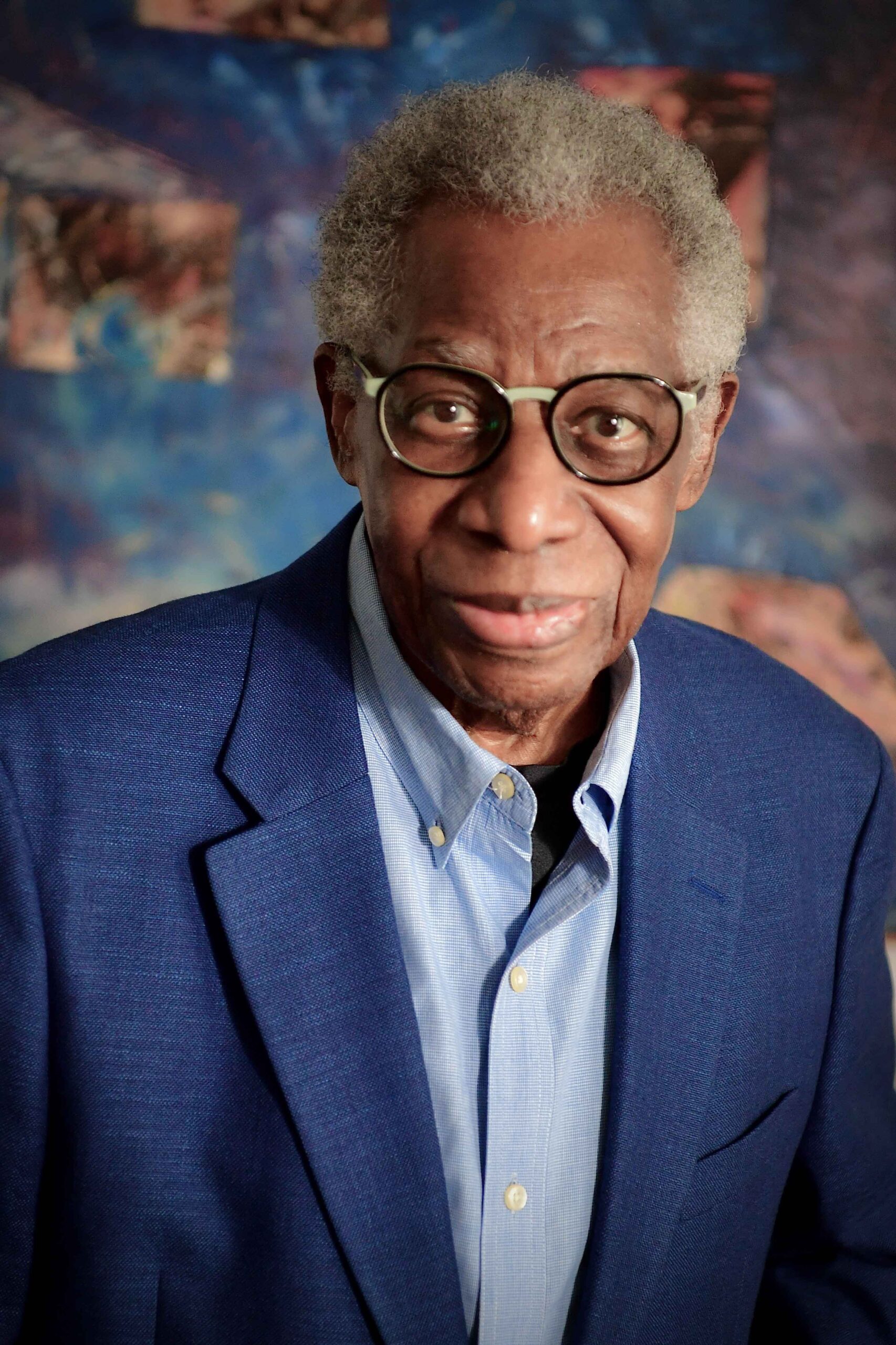 An older man with gray hair and glasses wears a blue blazer over a light blue shirt, smiling softly at the camera. The background is a mix of blue and brown abstract patterns.