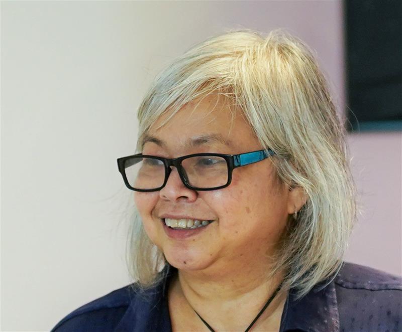 A woman with shoulder-length gray hair and glasses smiles. She is wearing a dark top and a black necklace, standing indoors with a soft, light-colored background.