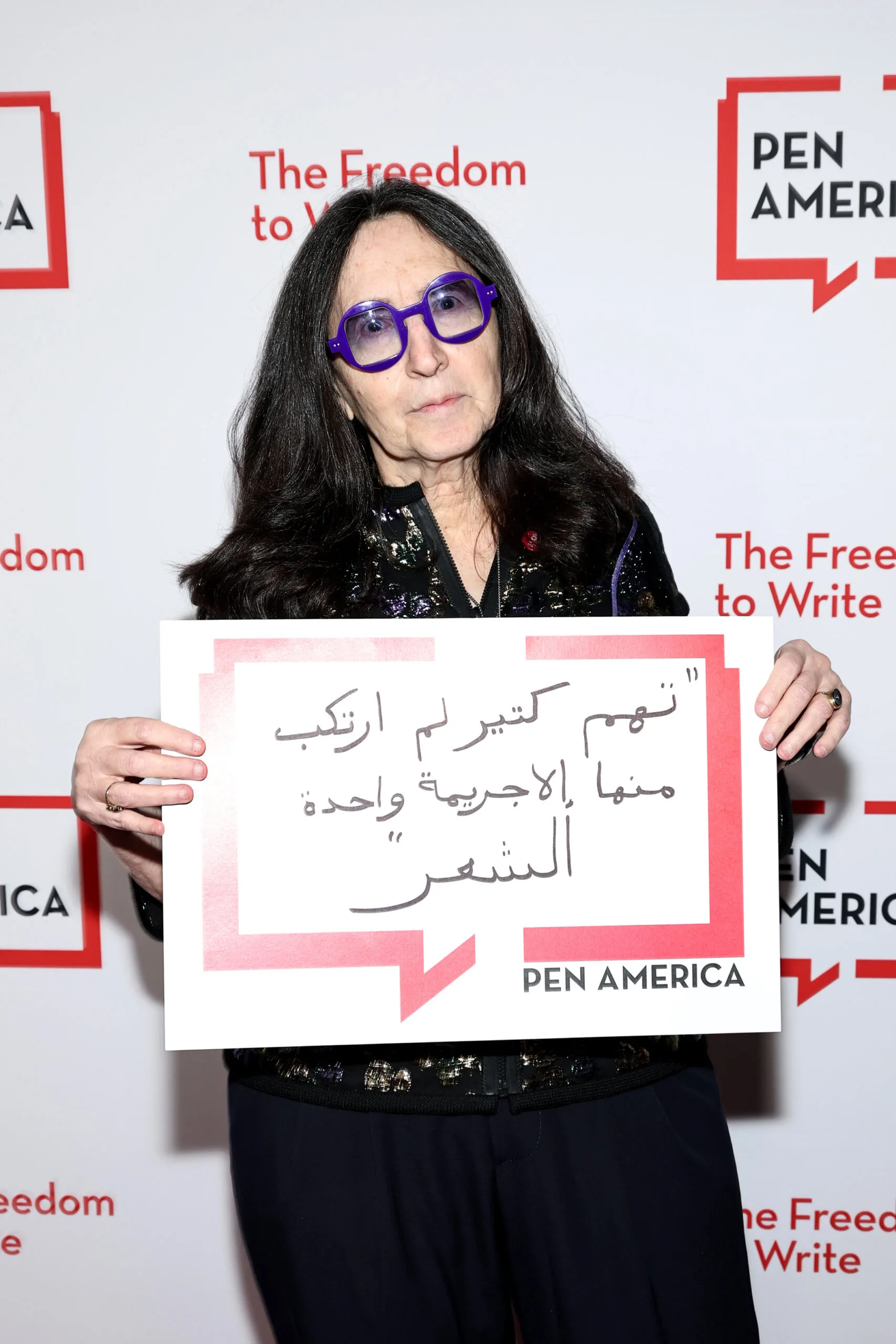 A woman with long dark hair and sunglasses holds a sign with Arabic text in front of a PEN America backdrop. The backdrop reads The Freedom to Write and PEN America in red and black.