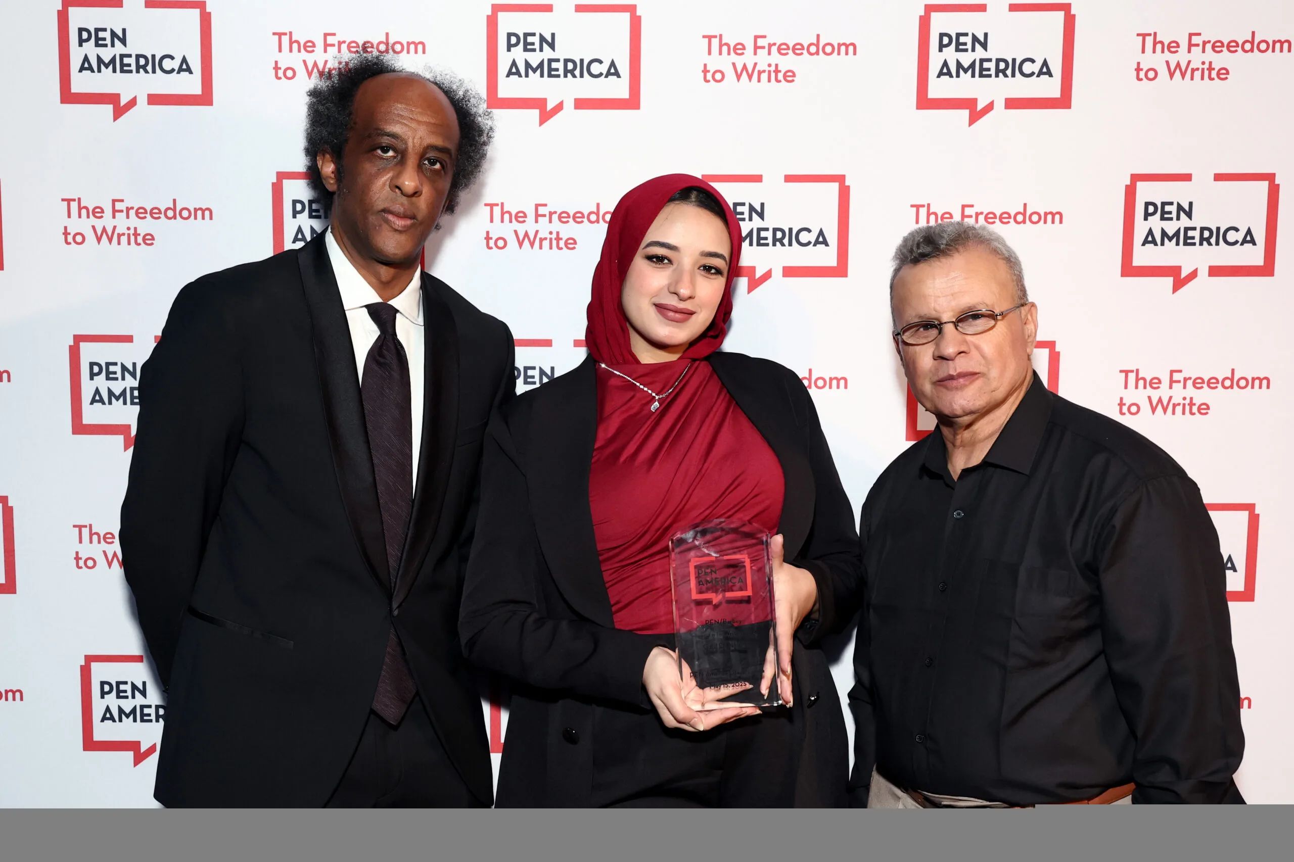Three people stand together at a PEN America event. The woman in the center, wearing a red hijab and holding an award, is flanked by two men. The background features the PEN America logo and “The Freedom to Write” slogan.