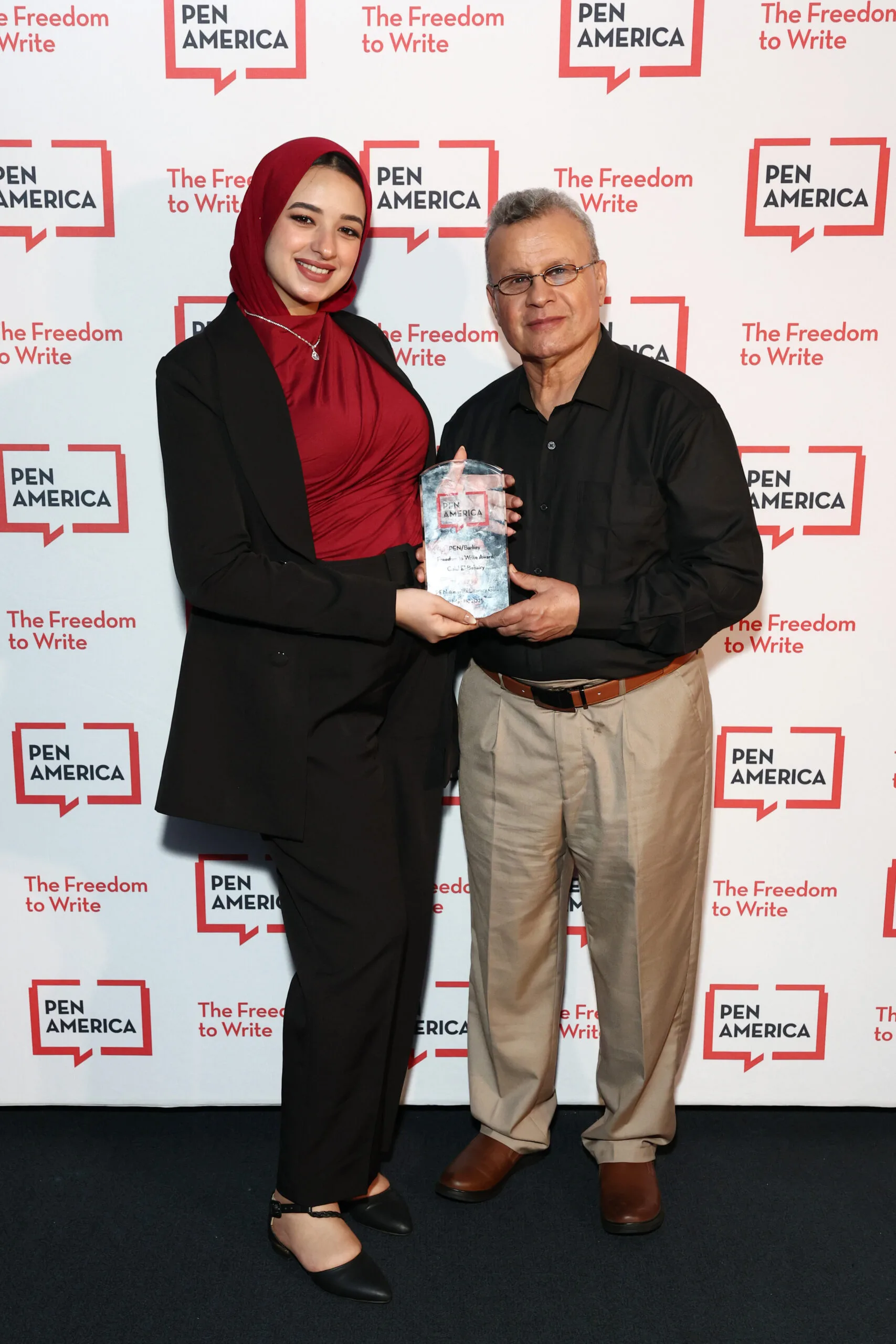 Two people stand in front of a PEN America backdrop. The woman on the left, wearing a red hijab and black outfit, holds an award with the man on the right, who is wearing glasses, a black shirt, and beige pants.
