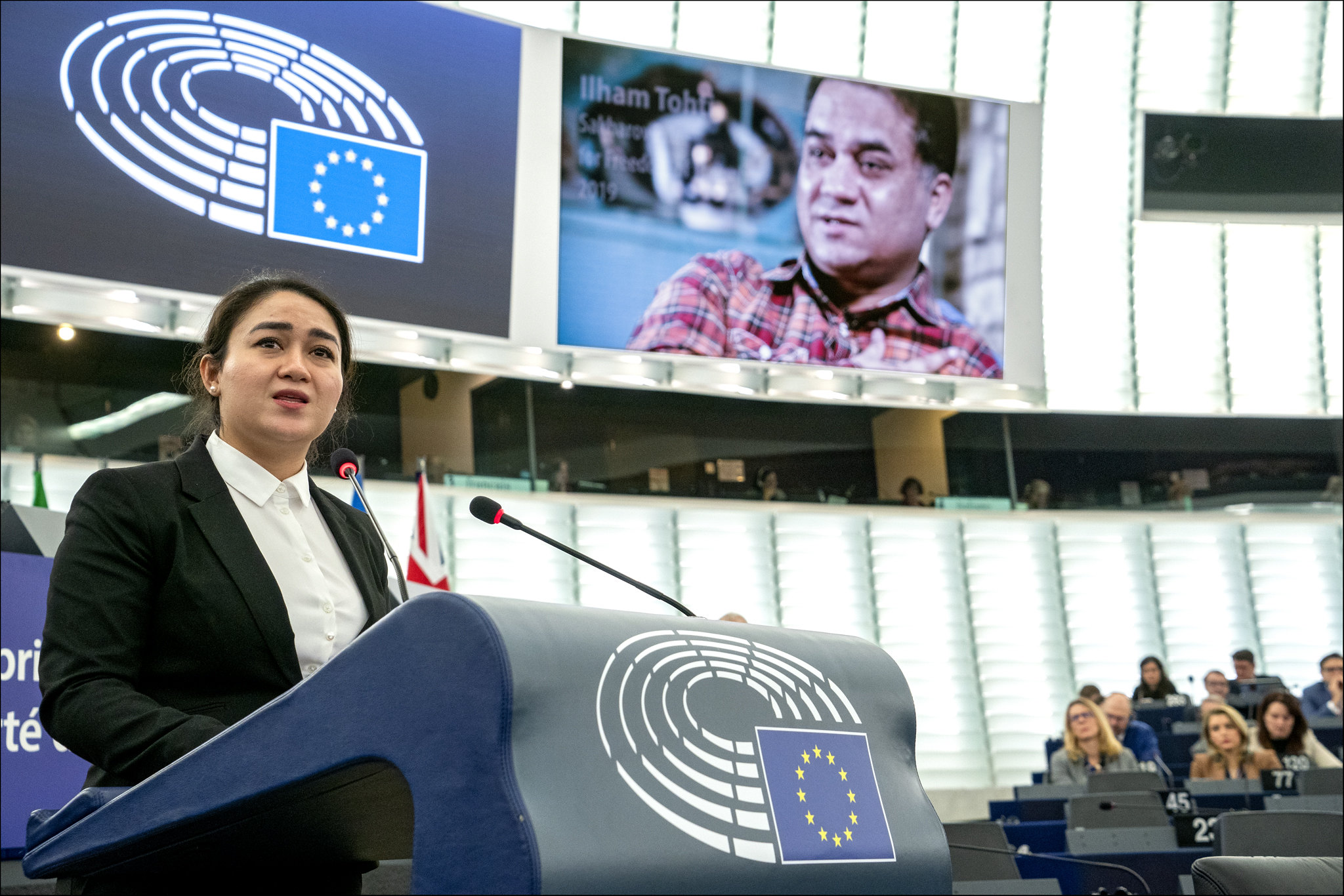 A woman speaks at a podium with the European Union emblem in a large assembly hall. Behind her, a screen displays a man’s portrait and the name Ilham Tohti. Seated audience members are visible in the background.