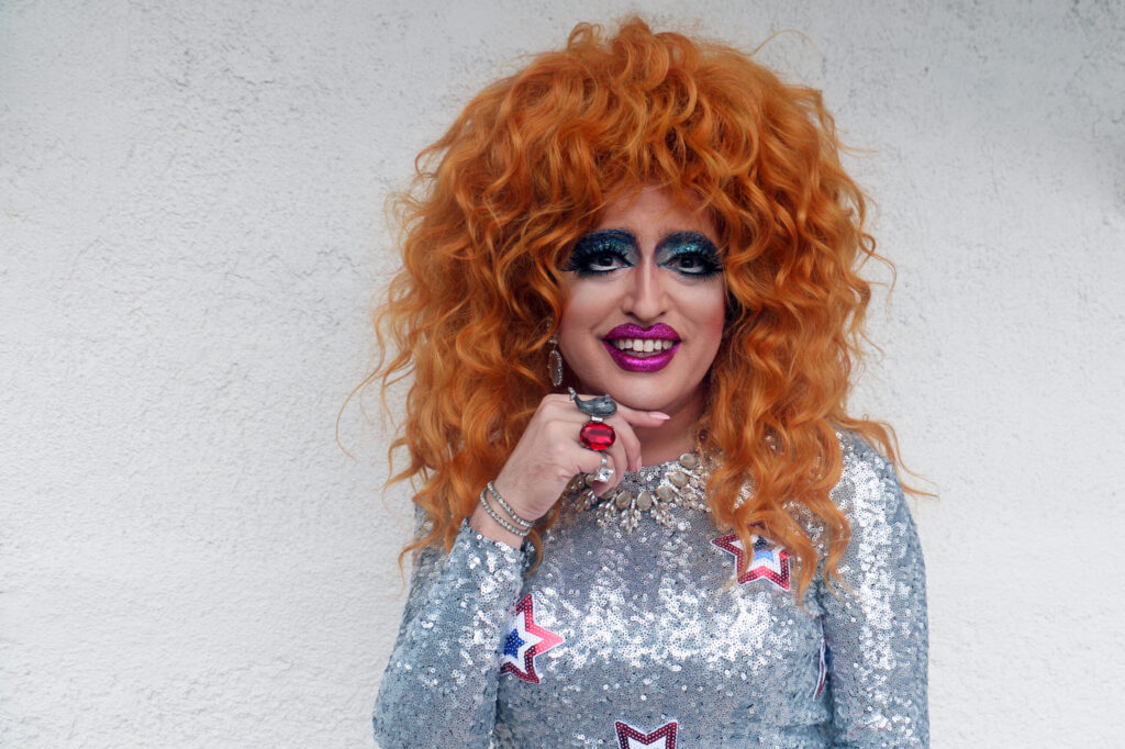 A person with voluminous orange curly hair, dramatic makeup, and bright pink lipstick wears a sparkly silver dress with star designs, a chunky necklace, and holds a hand with statement rings to their chin, standing against a plain light wall.