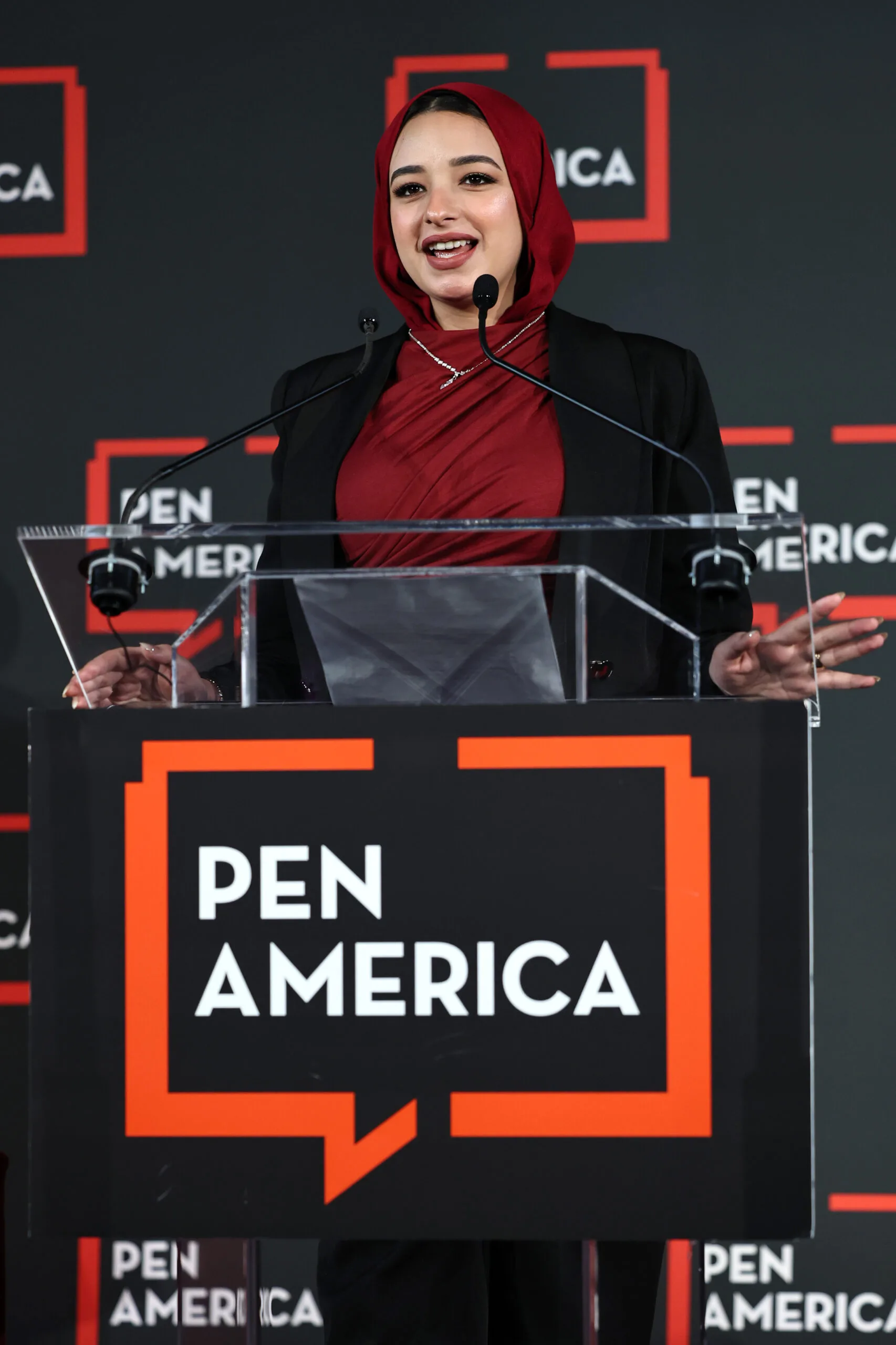 A woman in a red hijab and dark blazer speaks at a podium with a microphone. The podium and background display the PEN America logo in white and orange on black.