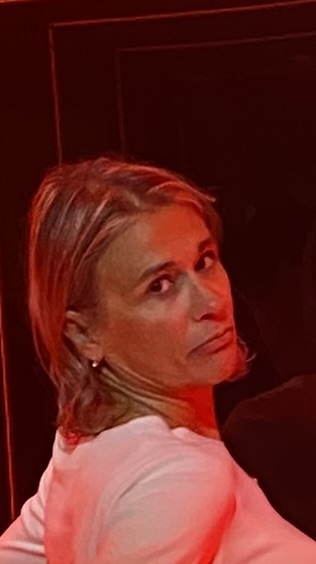 A person with short, light brown hair, wearing a white top, looks over their shoulder towards the camera with a neutral expression. The background is dark with reddish lighting.