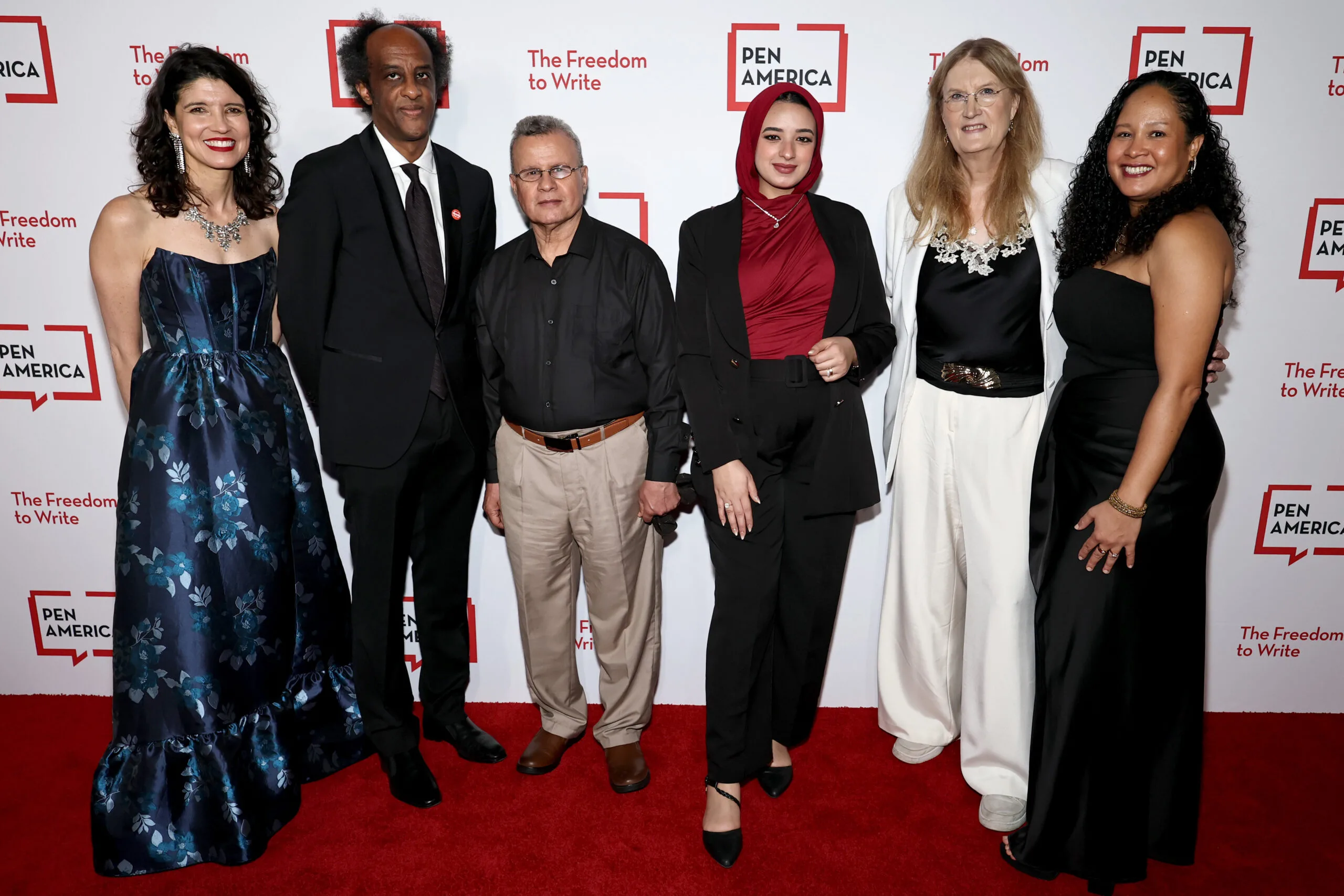 Six people stand together, smiling, on a red carpet in front of a PEN America backdrop that reads “The Freedom to Write.” The group is dressed in formal and semi-formal attire.