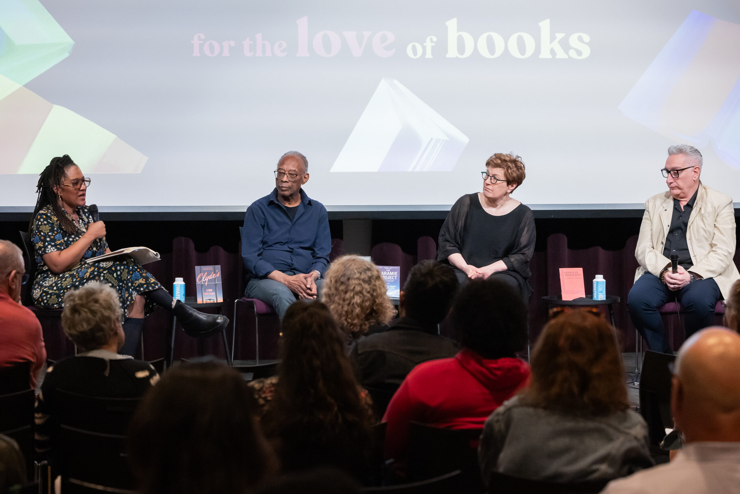Four people sit on stage in front of an audience, engaging in a panel discussion. A screen behind them reads For the Love of Books, and several titles, including works by Chimamanda Ngozi Adichie, are displayed on the table between them.