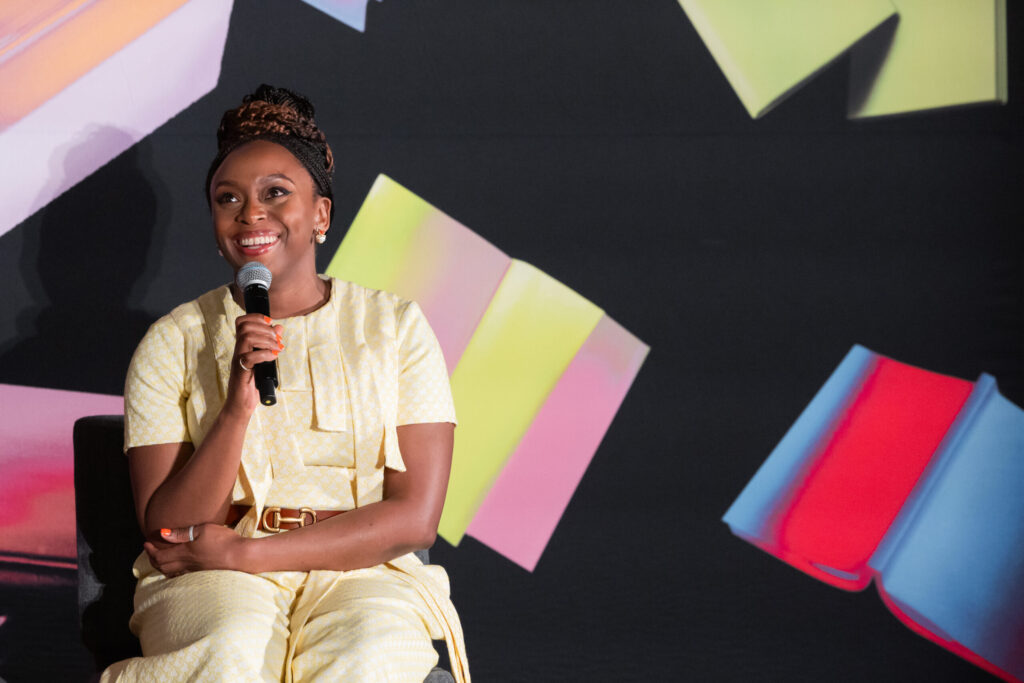 A woman in a light yellow outfit sits, smiling and holding a microphone, in front of a colorful background featuring large, blurred images of book covers.