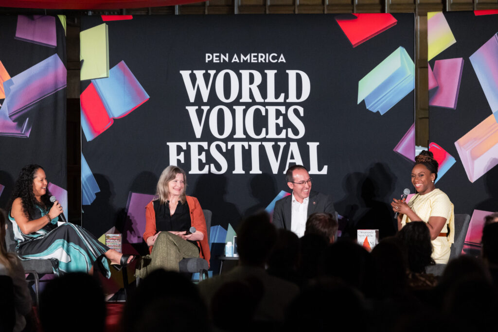 Four speakers sit on stage in front of a sign reading PEN America World Voices Festival, engaging in a lively panel discussion with an audience watching. Colorful book-like shapes decorate the background.