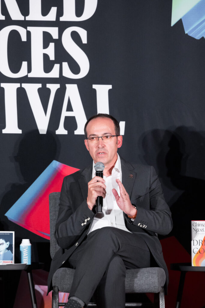A man in a suit sits on a chair, speaking into a microphone at an event. Behind him is a large sign with the words “RED FACES FESTIVAL” and colorful geometric shapes. Two books are visible on the table beside him.