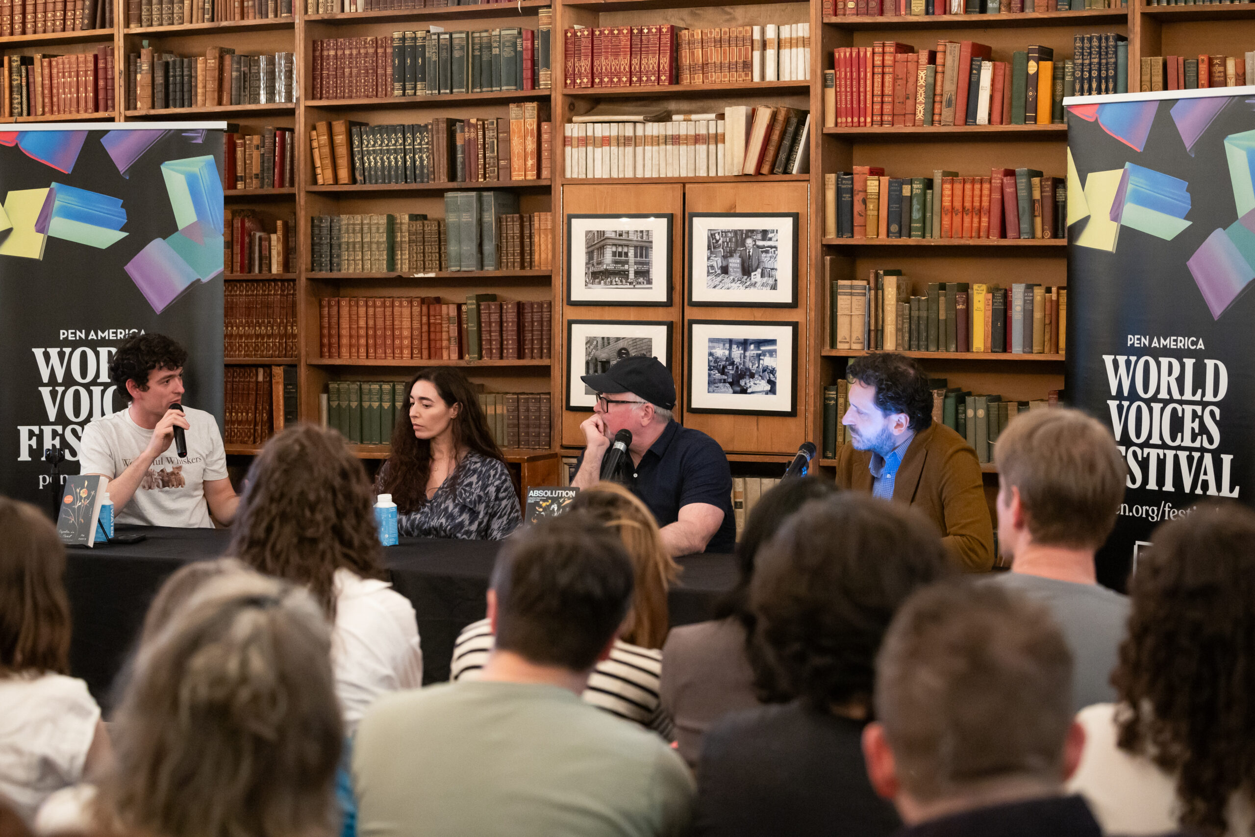 Four speakers sit at a table in front of an audience in a library setting, with bookshelves and PEN America World Voices Festival banners in the background. Two men and one woman are speaking into microphones.