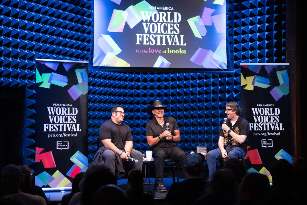 Three men sit on stage with microphones during a panel at the PEN America World Voices Festival. A screen and banners with the festival logo and colorful book graphics are in the background; audience members watch from the foreground.