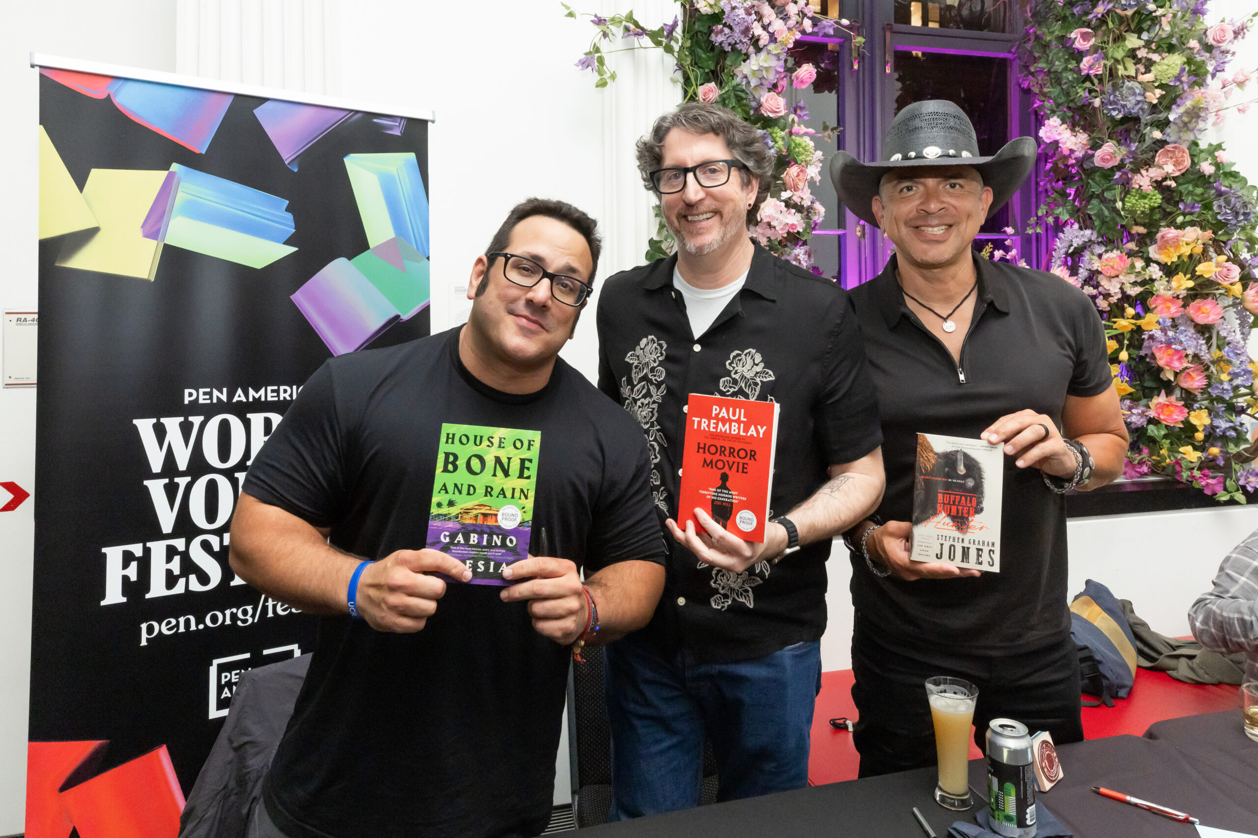 Three smiling men stand side by side at a table, each holding up a book. Behind them is a colorful PEN America World Voices Festival banner and a wall decorated with flowers and greenery.
