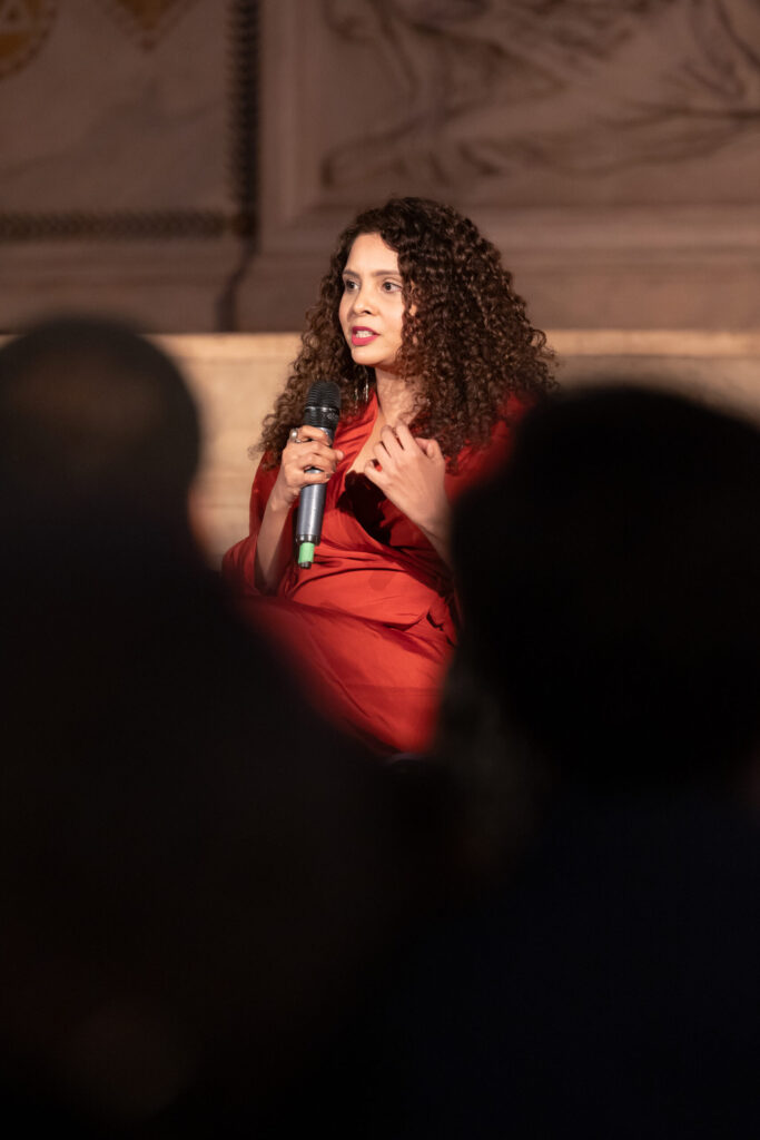 A woman with curly hair wearing a red outfit speaks into a microphone while seated on stage, with blurred audience members in the foreground.