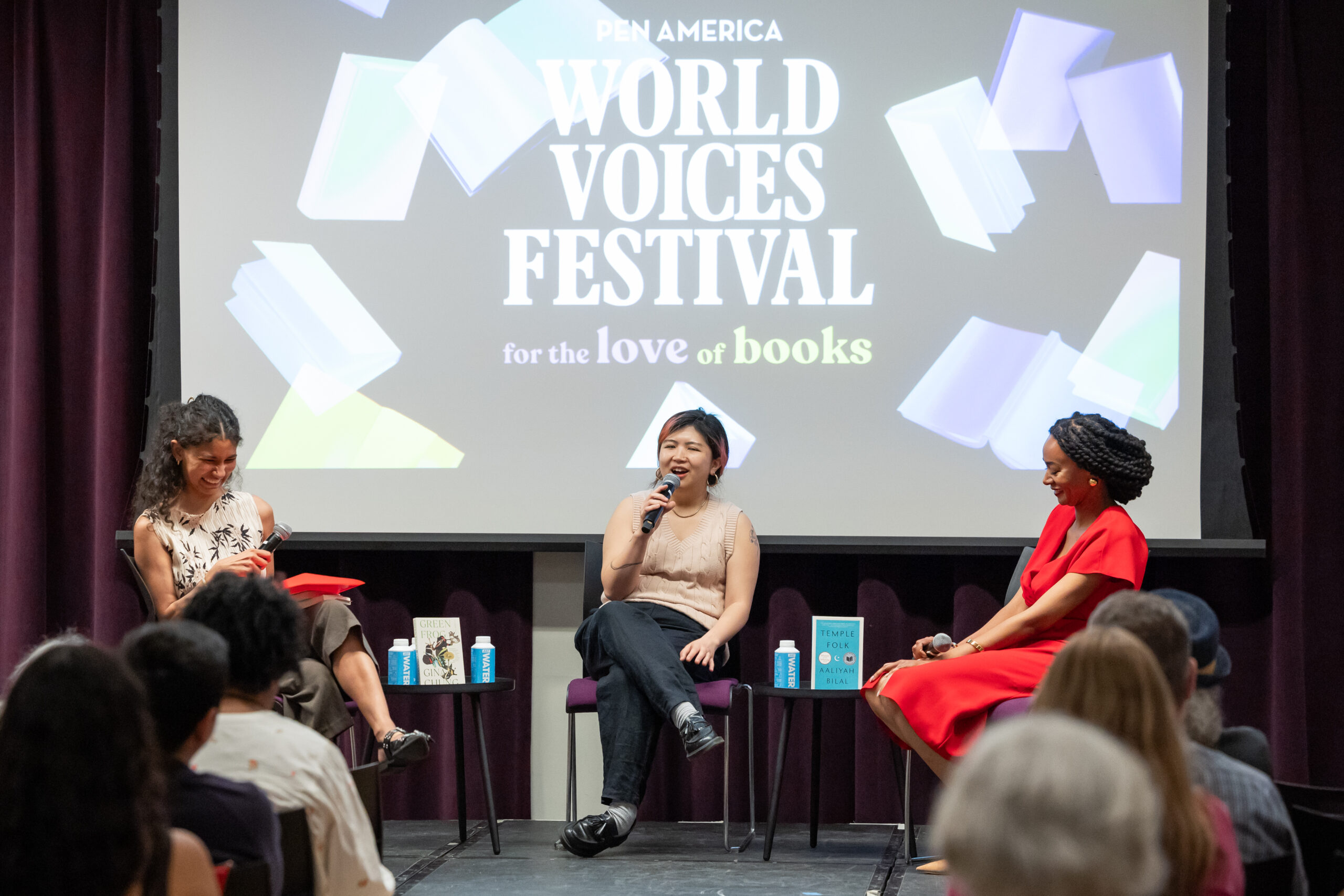 Three women sit onstage in front of a screen reading PEN America World Voices Festival for the love of books, engaging in a lively panel discussion before an audience. Books and water bottles are on the tables beside them.