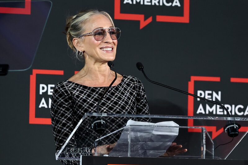 A woman with gray hair in a bun, wearing sunglasses and a black patterned dress, stands at a clear podium holding papers, smiling while speaking into microphones as she accepts the PEN America Literary Service Award. The background displays the PEN America logo.