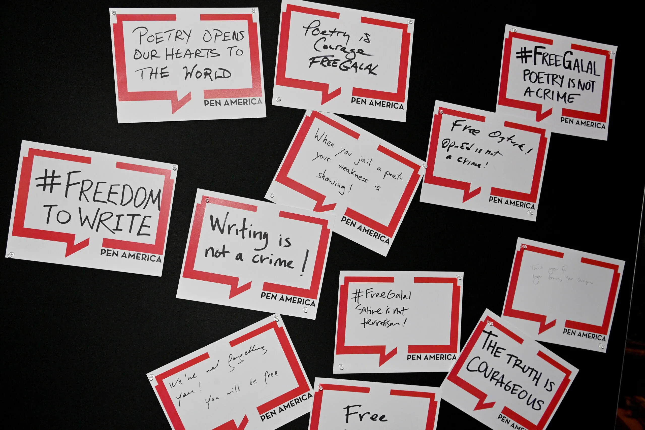 A collection of handwritten signs with messages like “#FREEDOMTOWRITE,” “Writing is not a crime!,” “Poetry opens our hearts to the world,” and “The truth is courageous,” displayed on a black background.