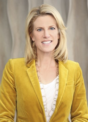 A woman with shoulder-length blonde hair wearing a mustard yellow blazer, a white top, and a gold chain necklace, smiling in front of a neutral, wavy-patterned background.