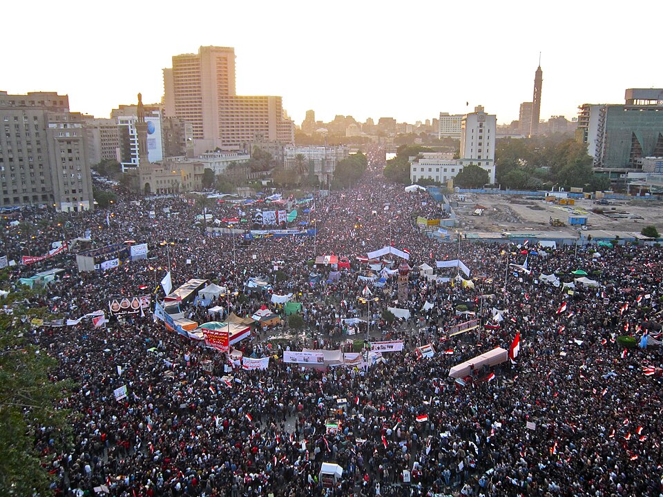 A large crowd gathers in a city square during daylight, holding flags and banners. Surrounding buildings and a tall tower are visible in the background. The scene suggests a protest or demonstration.