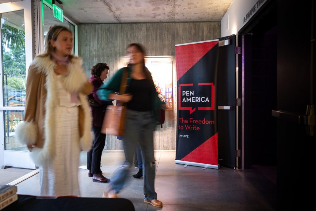 Two women walk past a PEN America sign that reads The Freedom to Write in a building hallway; the scene is slightly blurred, suggesting movement, with concrete walls and a lit exit sign in the background.