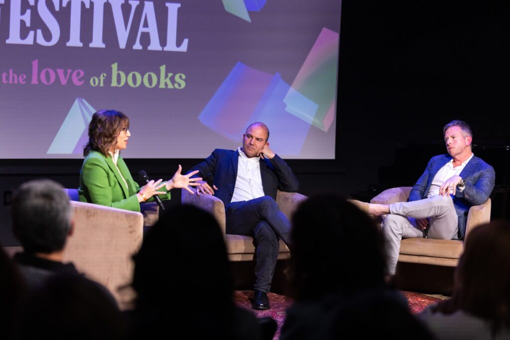 Three people sit on stage in armchairs, engaged in a panel discussion at a book festival. The woman on the left gestures while speaking, with two men listening. Projected text behind them reads, for the love of books.