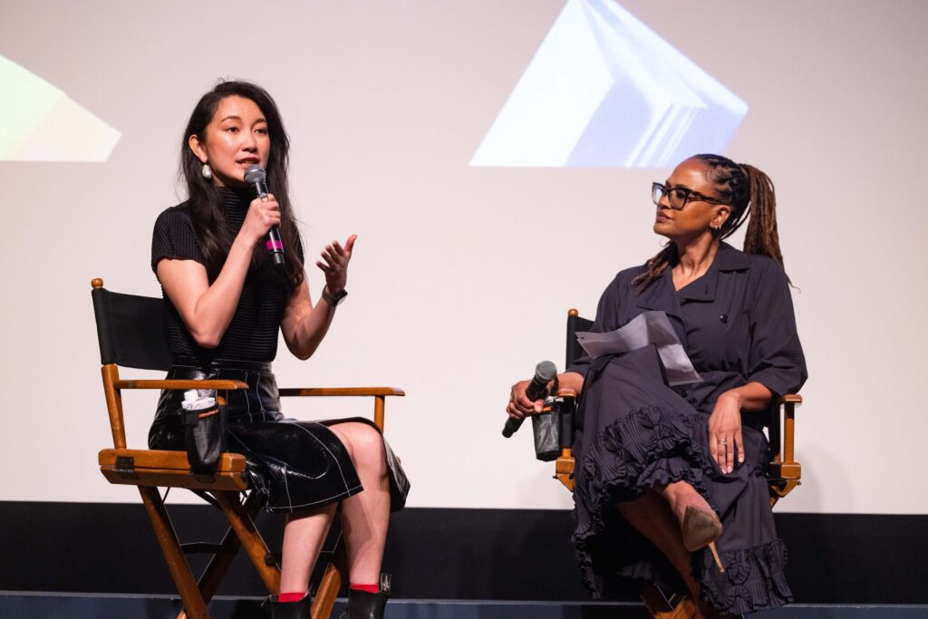 Two women sit on stage in director’s chairs having a discussion. One woman speaks into a microphone while gesturing, and the other listens attentively, holding a microphone and some papers. A projected image is visible behind them.