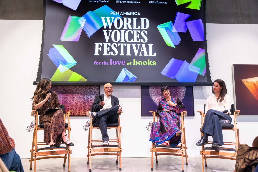 Four panelists sit on stage in front of a colorful PEN America World Voices Festival for the love of books sign, engaging in a discussion, with microphones and notes in hand.