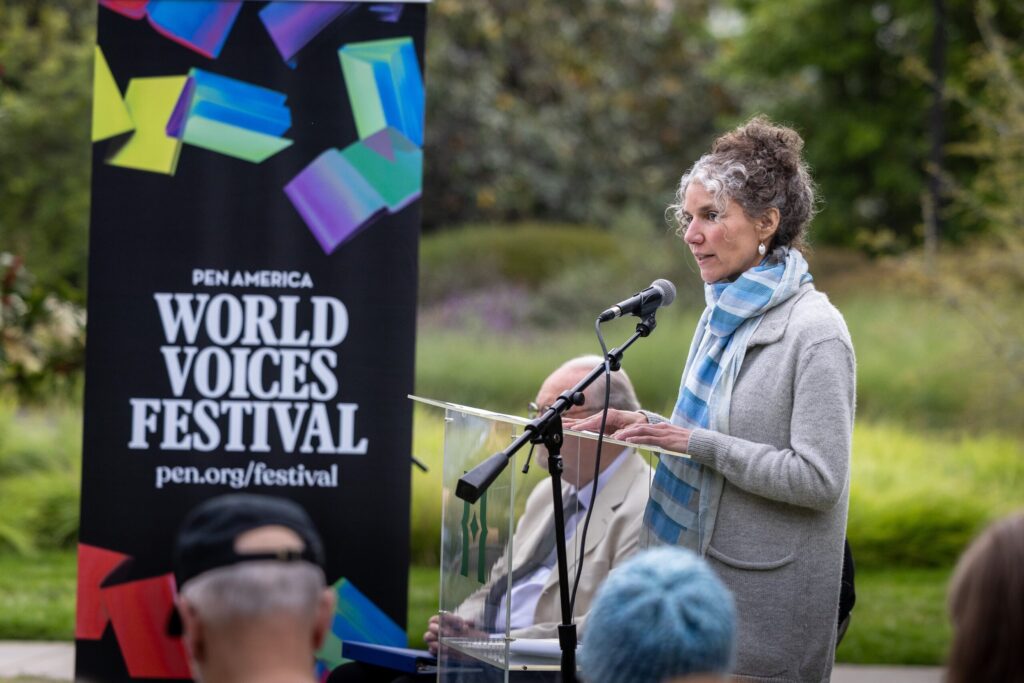 A woman in a gray sweater and blue scarf speaks at a podium outdoors during the PEN America World Voices Festival. A festival banner and seated audience members are visible in the background.