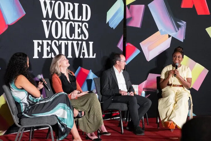 Four people sit on a stage with microphones in hand, engaging in a panel discussion at the World Voices Festival. Colorful geometric shapes decorate the backdrop behind them.