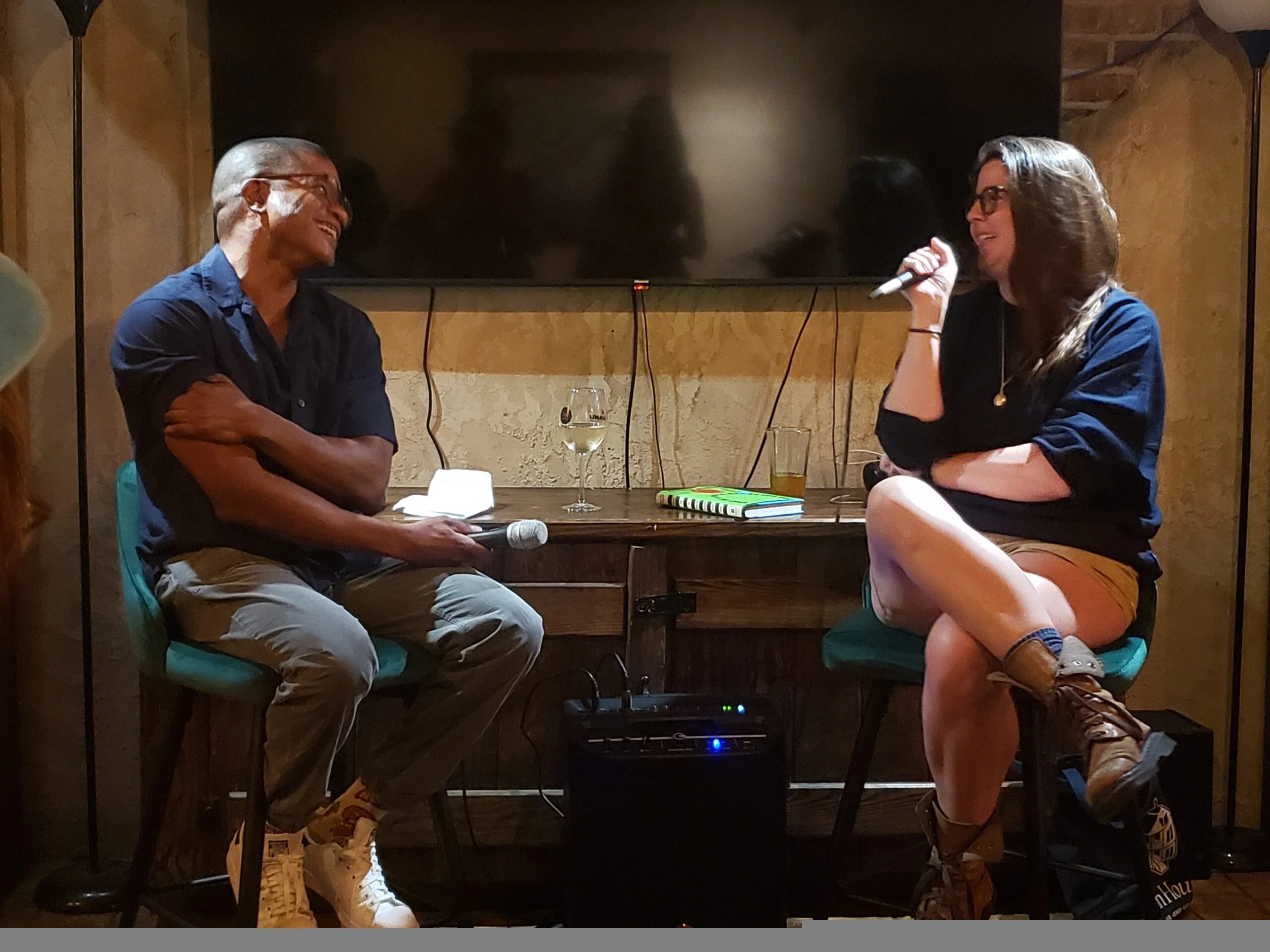 A man and a woman sit on stools, smiling and holding microphones during a conversation in a cozy room. A table with drinks and books is between them, and a TV is on the wall behind them.