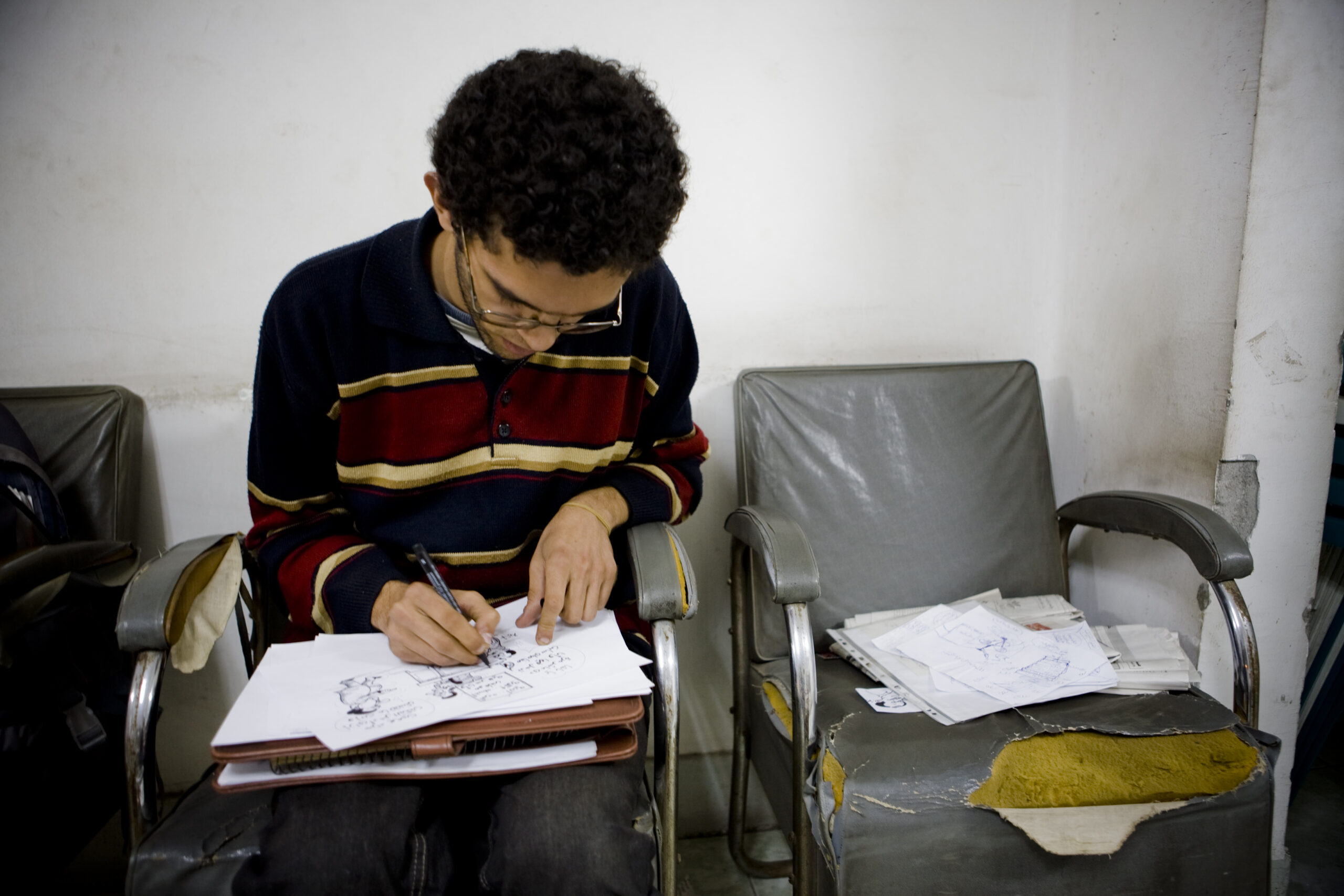 A person with curly hair sits and draws on a sketchpad, focused on their work. They wear glasses and a striped sweater. Next to them, a damaged gray chair holds scattered papers with drawings.