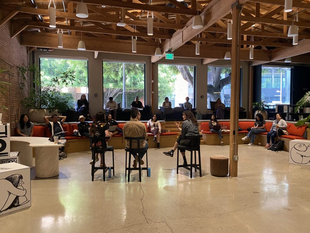 A group of people sit on stools and a long red couch in a spacious, modern room with wood beams, large windows, and greenery outside, attending what appears to be an informal meeting or discussion.