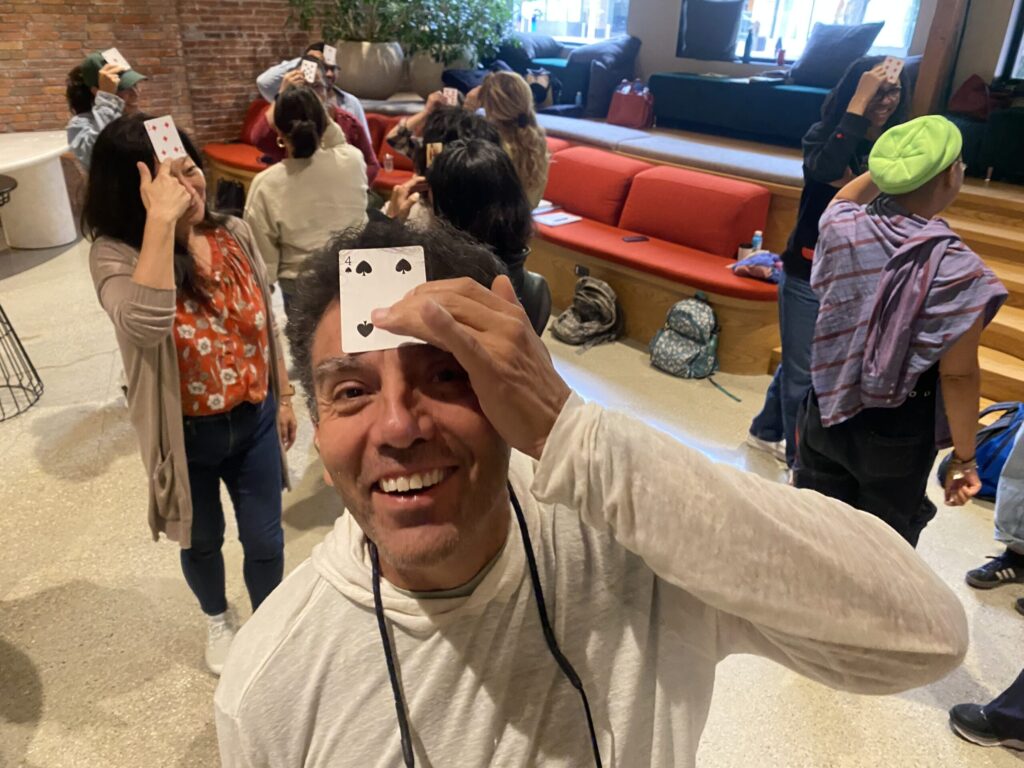 A smiling man holds a playing card to his forehead in a group setting, with others around him also holding cards to their foreheads in a casual indoor space.