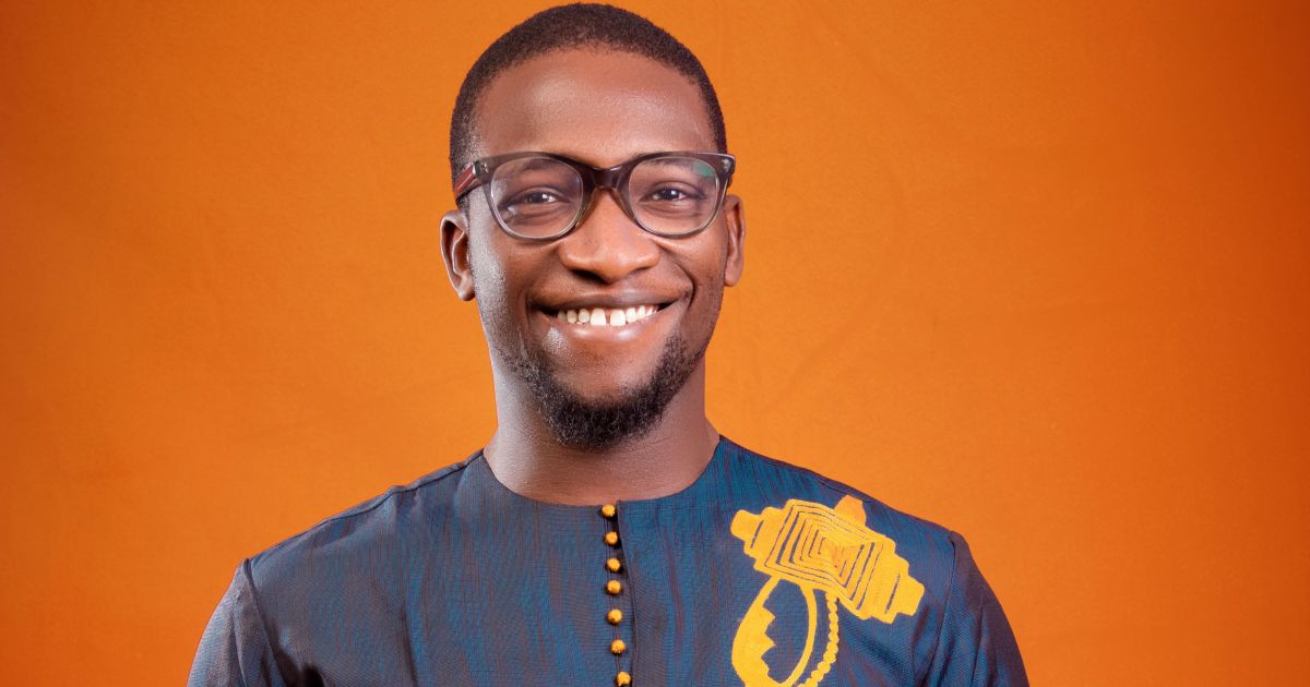 A man wearing glasses and a blue patterned shirt with yellow detailing smiles in front of an orange background.