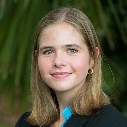A young woman with straight, shoulder-length blonde hair and green eyes, wearing a black blazer, light blue blouse, and gold hoop earrings, smiles softly in front of a blurred background of green foliage.