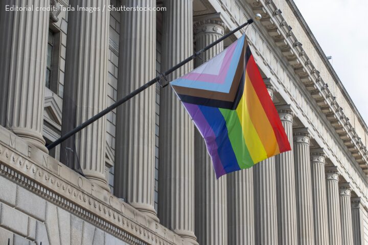 A Progress Pride flag flies on a pole outside a classical building with tall columns and stone details. The flag features rainbow colors plus chevrons in white, pink, blue, brown, and black.