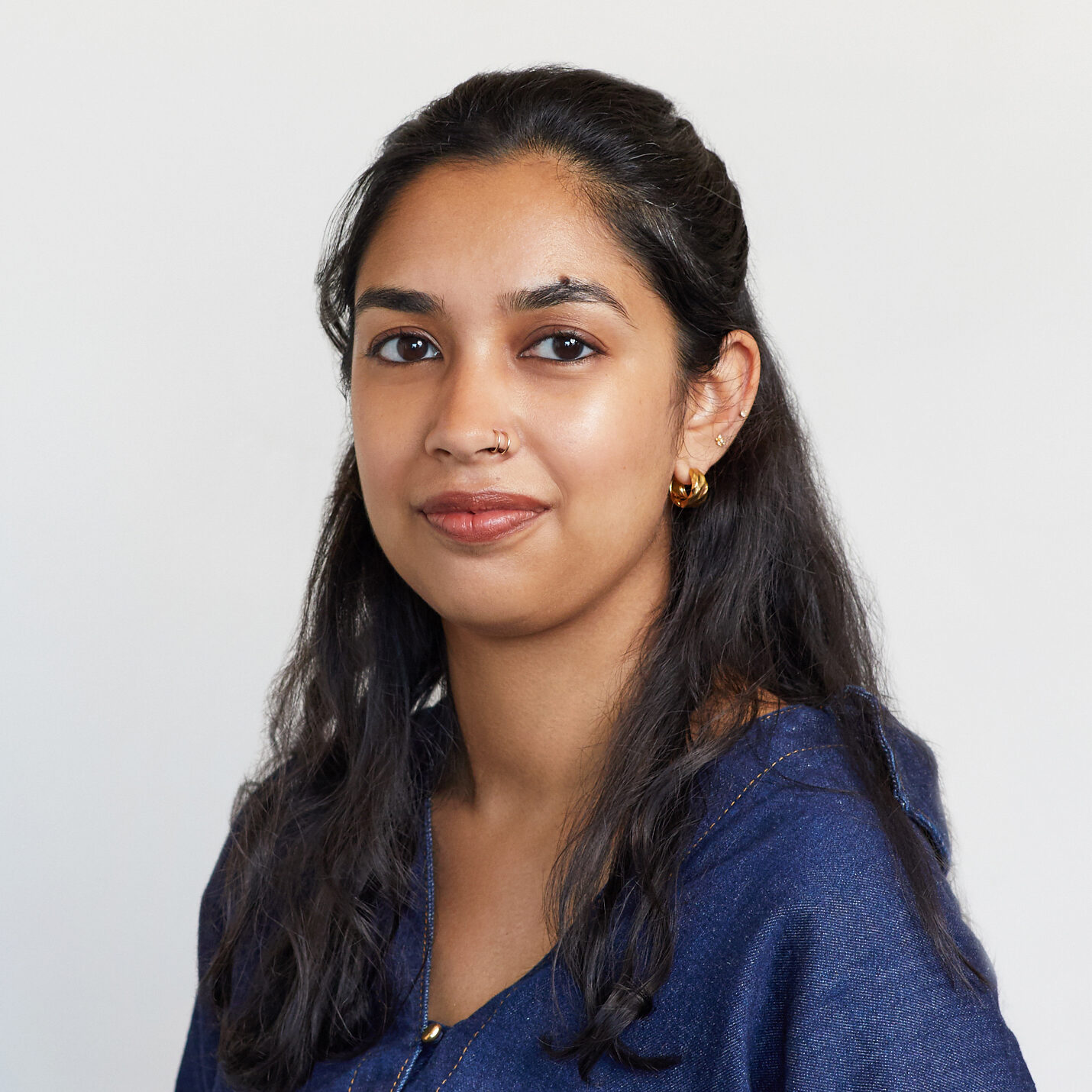 A woman with long dark hair wearing a blue top and gold hoop earrings looks at the camera with a neutral expression against a plain light background.