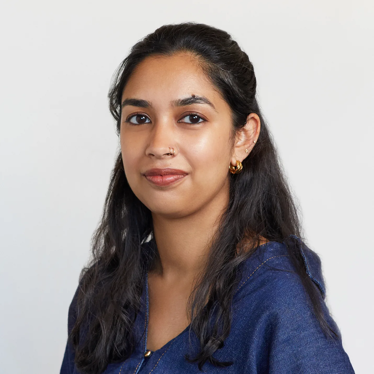 A woman with long dark hair wearing a blue top and gold hoop earrings looks at the camera with a neutral expression against a plain light background.