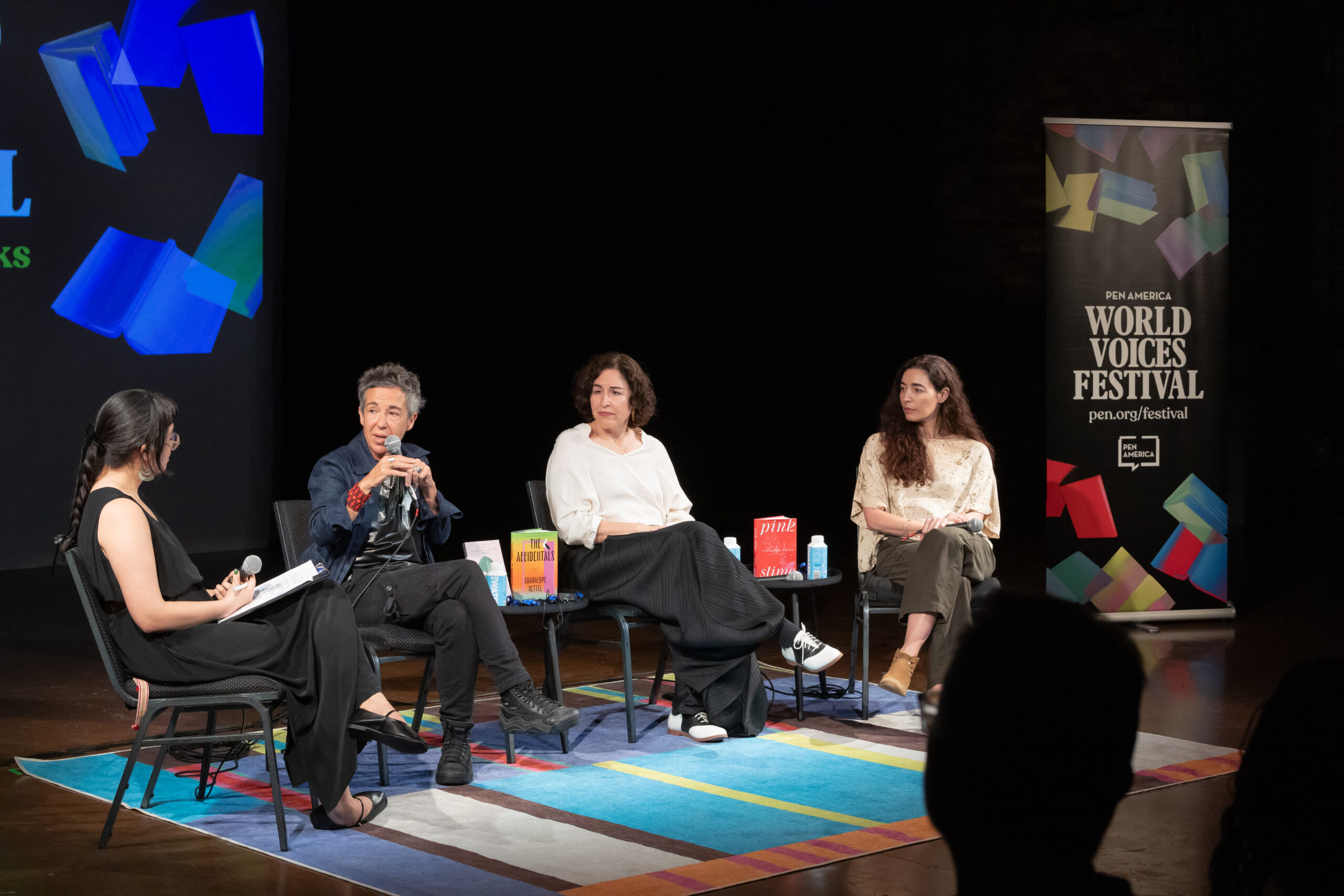 Four people sit on a stage having a panel discussion, each with a microphone. Books are placed in front of them. A PEN America World Voices Festival banner and colorful graphics are visible in the background.